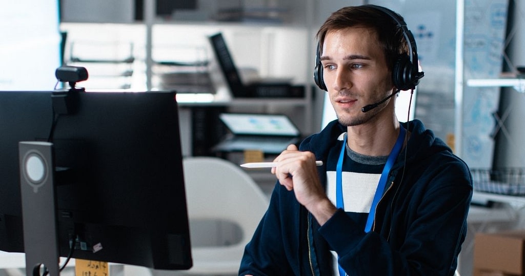 A man wearing headphones and holding a pen is working in a call centre and analysing data on a screen.