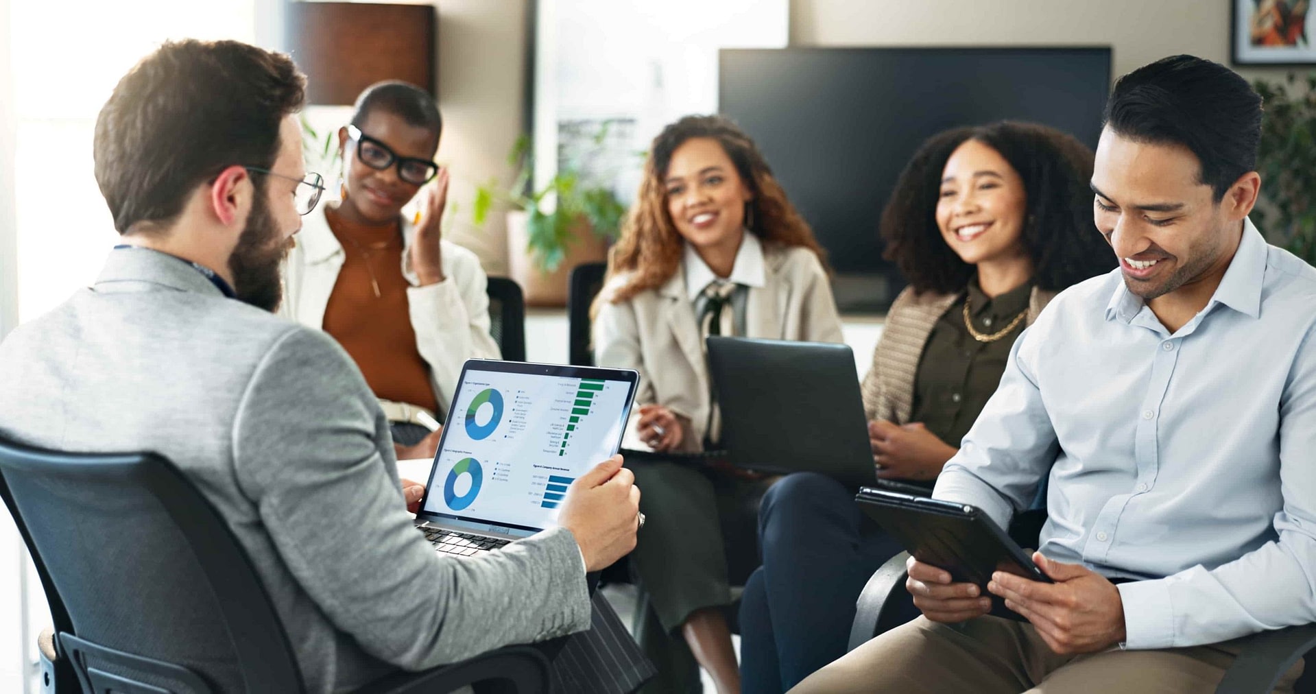 A seated man holding a laptop is presenting to a smiling team sitting in front of him.
