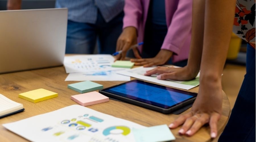 Work colleagues are collaborating around a table filled with charts, a laptop, and a tablet.