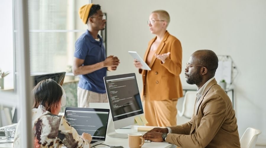 A team of colleagues working in an office, with two people at desks with computers and two standing and talking in the background.